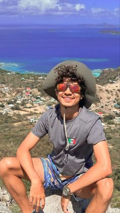 a young man sitting on top of a rock next to the ocean wearing a hat and sunglasses