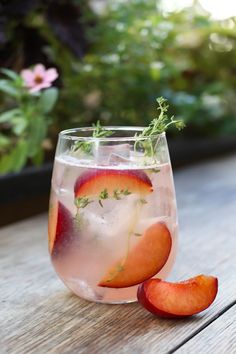 a glass filled with ice and sliced peaches on top of a wooden table next to potted plants