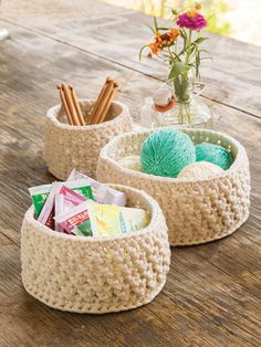 three baskets with yarn and knitting needles in them on a wooden table next to a vase filled with flowers