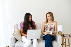 two women sitting on a couch with laptops in their hands and one holding a book