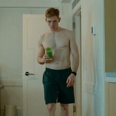 a shirtless man standing in a hallway holding a drink and looking at his cell phone