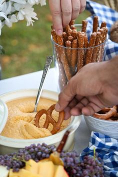 a person dipping pretzels into a bowl of dip with grapes and apples in the background