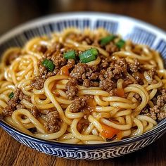 a close up of a bowl of noodles with meat and vegetables on top, sitting on a wooden table