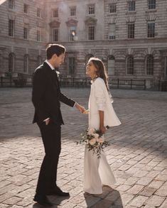 a bride and groom standing in front of an old building holding each other's hands