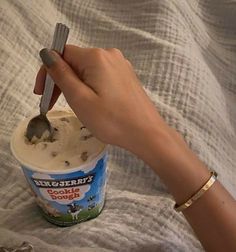 a hand holding a spoon over a cup of ice cream on top of a bed