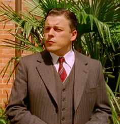 a man wearing a suit and tie standing in front of a brick wall with palm trees