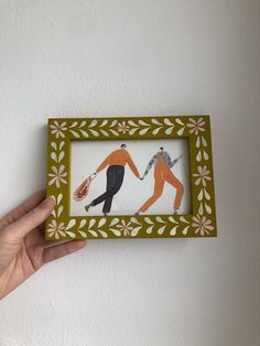 a hand holding a small framed artwork with two people dancing on the dancefloss