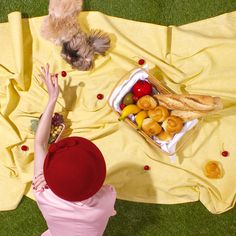 a woman in a pink dress and red hat standing next to a yellow blanket with food on it