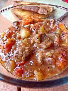 a glass bowl filled with stew and bread on top of a wooden table next to a cup of coffee