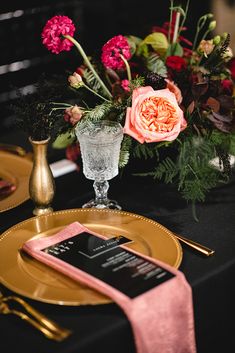 a table setting with pink flowers and gold place settings on black linens, silverware, and napkins