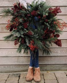 a woman is holding a wreath with red berries and greenery on the side of a building