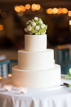 a three tiered white cake with flowers on top sits on a table in front of candles