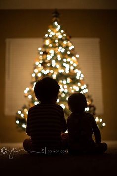 two children sitting in front of a christmas tree with the lights on it's windowsill