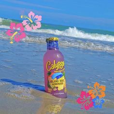 a pink bottle sitting on top of a sandy beach next to the ocean and flowers