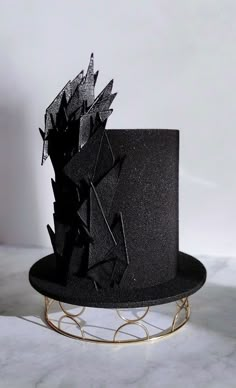 a black hat with an intricate design on top sits on a metal stand in front of a white wall