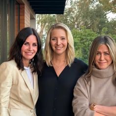 three women standing next to each other in front of a building with trees behind them