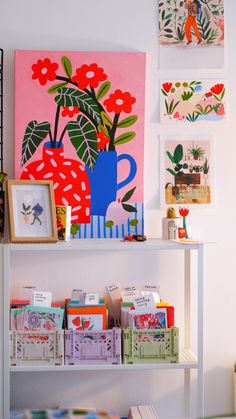 a shelf with books and pictures on it next to a wall filled with flowers, plants and paintings