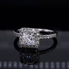 an engagement ring with a princess cut diamond surrounded by pave diamonds on a reflective surface