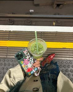 a person holding a cell phone with a drink in their hand on a train platform