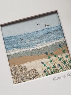 an embroidered picture frame with seagulls flying over the water and beach grass in front of it