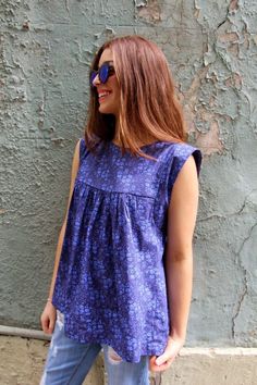 a woman standing in front of a wall wearing blue jeans and a purple top with flowers on it