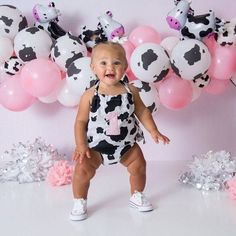 a baby girl standing in front of balloons and cow onesuits on a pink background