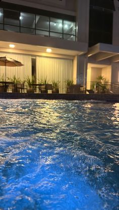 an empty swimming pool with chairs and umbrellas in front of a building at night