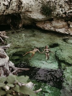 two people are floating in the water near some rocks and mossy vegetation, while holding hands