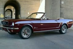 an old red mustang convertible parked in front of a concrete wall and doorway with the door open