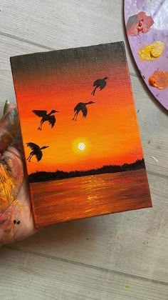 a person holding up a painting with birds flying over the water at sunset in front of them