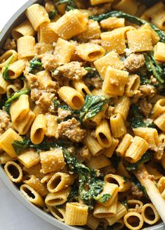 a pan filled with pasta and spinach on top of a white countertop next to a wooden spoon