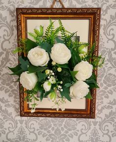 white roses and greenery are arranged in front of a gold framed wall hanging on the wall