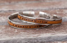 two bracelets with words on them sitting on a wooden surface, one has an orange band and the other is silver