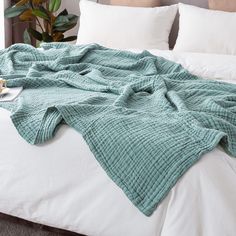 an unmade bed with white pillows and green blankets on it, next to a potted plant