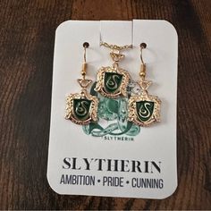 three pieces of green and gold jewelry sitting on top of a white card with the name slytherin