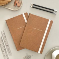 two brown notebooks sitting on top of a table next to a measuring tape and pencil