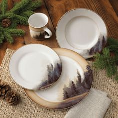 two white plates with trees painted on them next to pine cones and evergreen branches,