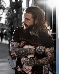 a man with long hair and tattoos on his arms is standing in front of a building