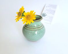 a green vase with yellow flowers in it on a table next to a piece of paper