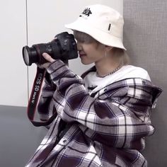 a person wearing a hat and holding a camera up to their face while standing next to a wall