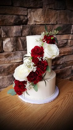 a wedding cake with red and white flowers on top is sitting on a table in front of a stone wall