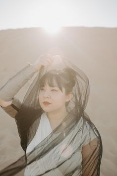 a woman standing in the sand with her hands on her head and wearing a veil over her head