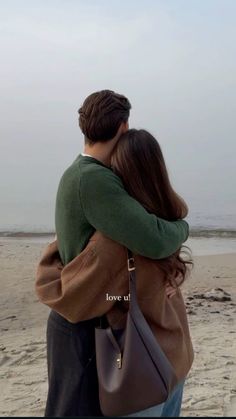 a man and woman hug on the beach while looking out at the ocean from behind them