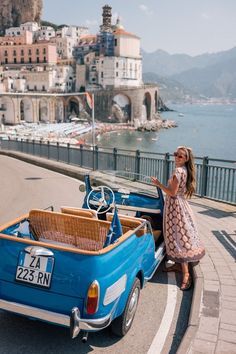 a woman standing next to an old blue car on the side of a road with buildings in the background