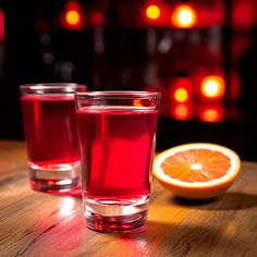 two glasses of red liquid sitting on top of a wooden table next to an orange