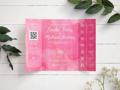 a pink watercolor wedding card with white lettering on it and greenery next to it