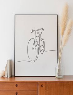 a vase with some dry grass in it next to a framed bicycle print on a dresser