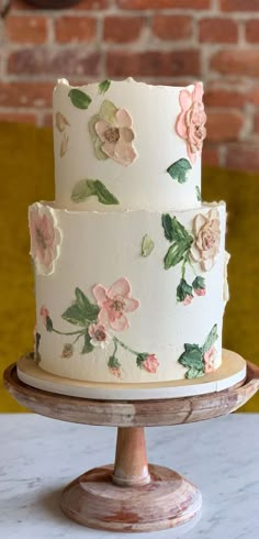 a white cake with pink flowers on it sitting on a wooden stand in front of a brick wall