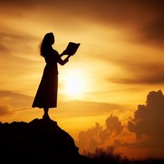 a woman standing on top of a hill holding a book in her hands while the sun is setting