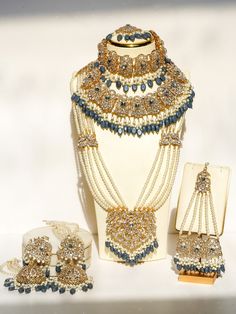 Experience the ultimate luxury with the Nazanin Gold Plated Bridal Set, a meticulously crafted 6-piece collection designed to captivate and charm. This opulent bridal set includes oversized jhumka earrings that are 4.3 inches long with attached ear chains, a resplendent choker necklace, a timeless classic necklace, a stunning long necklace, a graceful jhoomer, and a majestic maang tikka, all adorned with exquisite gold plating, shimmering white zircon stones, and captivating pantone blue beads t Blue Luxury Wedding Sets, Luxury Blue Wedding Sets, Elegant Meenakari Sets For Ceremonial Occasions, Elegant Blue Bridal Necklace With Intricate Design, Blue Bollywood Bridal Necklace For Wedding, Blue Hand Set Bridal Necklace For Reception, Elegant Bridal Sets With Zari Work For Ceremonial, Elegant Bridal Sets With Zari Work For Ceremonial Occasion, Blue Meenakari Sets For Wedding